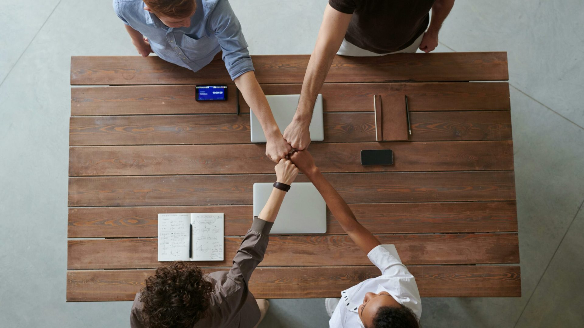 A diverse group of professionals engaging in a teamwork celebration with a fist bump over a wooden table indoors.